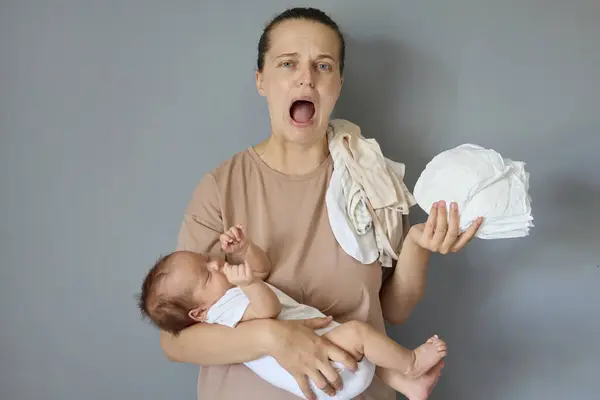 stock image Despair helpless Caucasian young woman wearing beige T-shirt standing with newborn baby isolated over gray background screaming loud and crying being tired needs help with her infant child