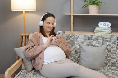 Gülümseyen kahverengi saçlı beyaz hamile kadın evdeki koltukta oturuyor ve akıllı telefonu kulaklıkla veya ses podcasıyla müzik dinlemek için kullanıyor.