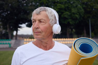 Beyaz tişörtlü gri saçlı sporcular stadyumda kulaklıklarıyla yoga minderlerini tutuyor gözlerini kaçırıyor açık hava antrenmanı için kanepede müzik dinliyor.