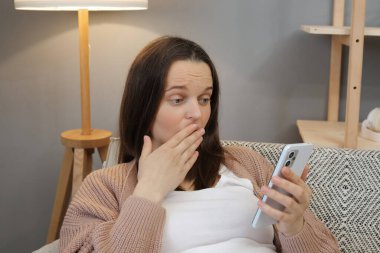 Şaşırmış genç hamile bir kadın, evindeki rahat oturma odasında akıllı telefonuyla kanepede rahatlarken sosyal ağda web sayfaları okurken şok edici içerikler görüyor.