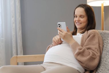 Neşeli, mutlu, hamile bir kadın akıllı telefonu web sayfalarını karıştırmak için kullanıyor. Evde cep telefonuyla gezen hamile anneler için forumlar hakkında bilgi okuyor.