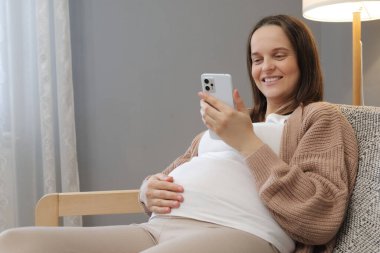 Akıllı telefonlu memnun hamile bir kadın evinde koltukta oturup interneti kullanıyor. Cep telefonu beklentisi olan annesi cep telefonu uygulamasında selfie mesajlaşması yapıyor.