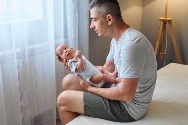 Babalık anıydı. Yetişkin, beyaz, yakışıklı baba yatak odasında iç tarafta oturuyor. Yeni doğmuş erkek çocuğu kucağında.