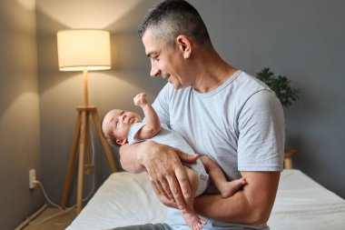 Neşeli beyaz adam, genç baba, kızına ya da oğluna şefkatle sarılıyor ve yeni doğmuş bebeğine sevgi ve sıcak bir gülümsemeyle bakıyor.