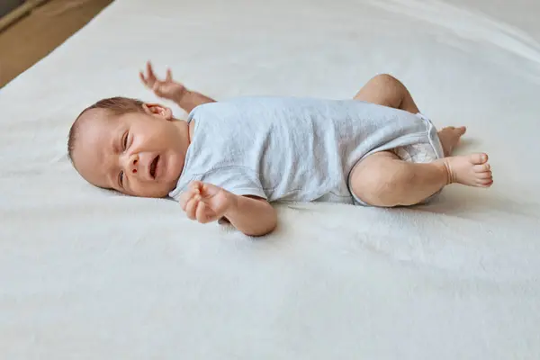 Stock image Newborn baby wearing bodysuit crying while laying on white sheet having childish tantrums suffering colic and abdominal pain infants needs medicine help or mother attention