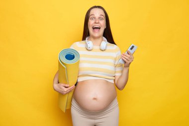 Aşırı neşeli kahverengi saçlı hamile kadın, cep telefonu çığlıkları atarak heyecan verici bir şekilde spor uygulaması indiriyor. Elinde karemat ve akıllı telefon, sarı arka planda tek başına duruyor.