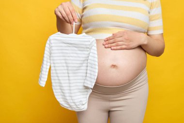 Çıplak karnıyla tanınmayan hamile bir kadın yeni doğan bebek için kıyafet alıyor. Doğuma hazırlanıyor ve annelik sarı arka planda tek başına duruyor.