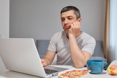 Sıkıcı, Kafkasyalı, gri tişörtlü esmer adam evde otururken dizüstü bilgisayarda çalışıyor bilgisayarla uzaktan kumandayla kafasını kaldırıp monitöre bakıyor.