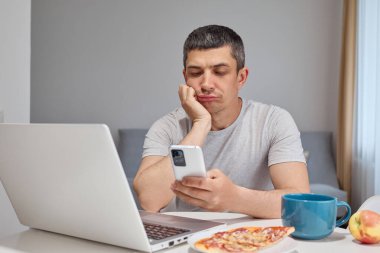 Düşünceli, sakin, günlük kıyafetler giyen, akıllı telefon ve dizüstü bilgisayar kullanan, uzaktan kumandalı bilgisayarını kullanarak cep telefonuyla kayan, ara verip evde oturup sıkıcı web sayfalarını karıştıran bir adam.