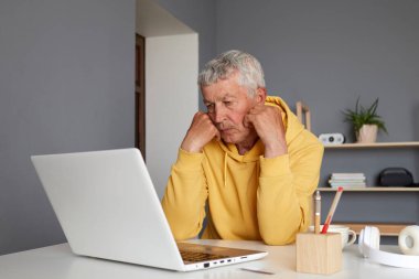 Sıkıcı Kafkasyalı olgun bir adam internette çalışıyor dizüstü bilgisayarda Webinar Podcast izliyor çalışma masasında oturuyor sarı kapüşonlu giyen son sınıf öğrencisi bilgisayar ekranına bakıyor.