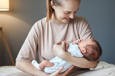 Gülümseyen, pozitif, genç, yetişkin bir anne, küçük oğlu ya da kızıyla birlikte yatak odasında oturuyor.