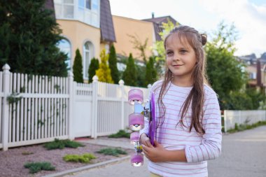 Gündelik giyinmiş, beyaz kadın çocuk sokakta kaykay numaraları yapıyor. Şehirde para tahtasını kucaklıyor ve büyüleyici bir gülümsemeyle başka tarafa bakıyor.