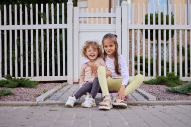 İki kız kardeş asfalt yolda otururken sarılıyor çocuklar sokakta eğleniyor dalgalı saçlı küçük çocuk kameraya gülümseyen büyük çocukla gülüyor