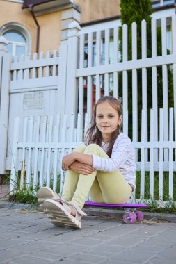 Cute Caucasian little girl playing skateboard or surf skate in city street learning skills looking at camera enjoying sport activity on her summer holiday clipart