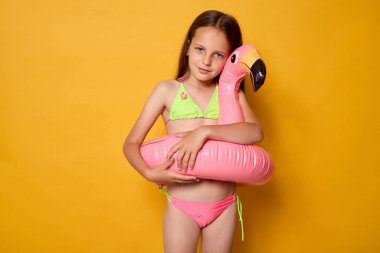 Kahverengi saçlı beyaz yüzücü kız flamingoyu kucaklıyor havuzda ya da nehirde yüzüyor sarı arka planda izole bir şekilde poz veren kameraya bakıyor.