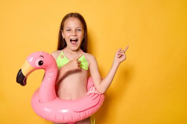 Mayo giymiş, belinde flamingo yüzükler olan neşeli küçük bir kız. Sarı duvara yaslanmış. Reklam amaçlı reklam merkezleri için fotokopi alanını gösteriyor.