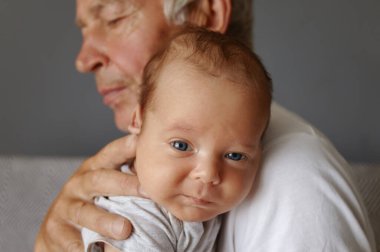 Vücut giysisi giymiş sevimli, sağlıklı bir bebek. Büyükbabanın kollarında huzur içinde yatıyor. Küçük bebek dedenin ellerinde uyuyor.