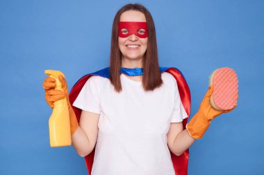 Gülümseyen, maskeli, pelerinli, deterjan tutan, sünger temizleyen ve mavi arka planda tek başına duran neşeli kadın süper kahraman.