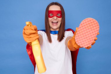 Maske takıp, pelerin takarak, temizlik spreyi sıkarak ev işlerini ve hijyen işlerini yürüterek mavi arka planda kalan son derece mutlu bir ev hanımı.