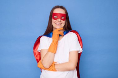 Kırmızı süper kahraman giymiş, kendine güvenen beyaz kadın temizlikçi ev temizliğine hazır. Mavi arka planda tek başına ayakta duran kameraya bakıyor.