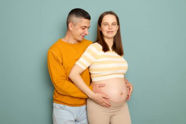 Romantik anne babalar sıcak bir anın tadını çıkarıyorlar. Bebek kocasının güzel hamile karısına sarılıp turkuaz arka planda çıplak karnına dokunmasını bekliyorlar.