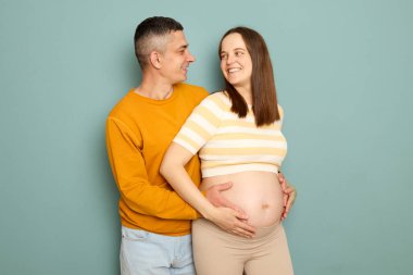 Sevimli, sevimli, sevgi dolu bir aile bebek bekleyen, turkuaz bir arka planda el ele tutuşan, sevgi dolu kocasına bakan, gülümseyen bir kadın.