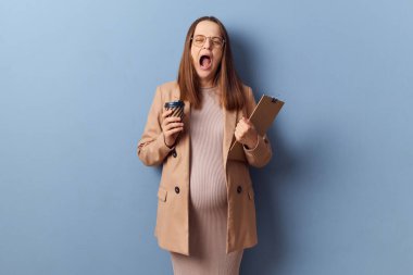 Yorgun, esnemiş kahverengi saçlı, gözlüklü, büyük göbekli, mavi arka planda izole edilmiş, kağıt klasörde saklanan, tek kullanımlık kahve içen bir iş kadınının çalışmak için enerjiye ihtiyacı var.