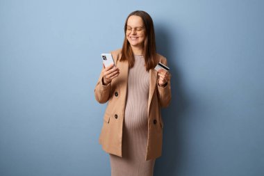 Çevrimiçi işlem. Banka teknolojisi. Bej ceket ve elbise giymiş gülümseyen hamile bir kadın mavi duvara yaslanmış cep telefonuyla eşyaları alıyor ve internetten ödeme yapıyor.