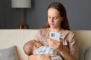Kahverengi saçlı, beyaz saçlı, genç bir anne. Kanepede oturup internet sayfalarını karıştırırken küçük kızı ya da oğlunu elinde tutuyor.