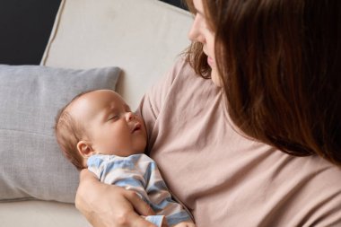 Çizgili elbiseli küçük bebeğin yakın çekimi. Anne yeni doğmuş bebeğini sallıyor. Oturma odasında kanepede oturuyor ve kucağında uyuyan bir çocuk ya da kız çocuğu var.