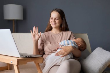 Bej tişört giymiş, gülümseyen bir kadın bebekle kanepede oturuyor ve internette konuşurken onu sallıyor. İnternetin tadını çıkarıyor, selamlama hareketi yapıyor.