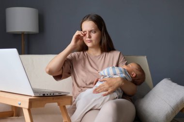 Yorgun bir anne evdeki kanepede oturup dizüstü bilgisayarını uzaktan çalışmak için kullanırken bebeği de elinde uyuyordu teknolojiyi ebeveynlikle harmanlıyordu acı hissediyordu.