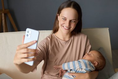 Genç, güzel, güler yüzlü bir anne akıllı telefonuyla yeni doğmuş bebeğiyle evde otururken annelik blogunda içerik oluşturarak selfie çekiyor.