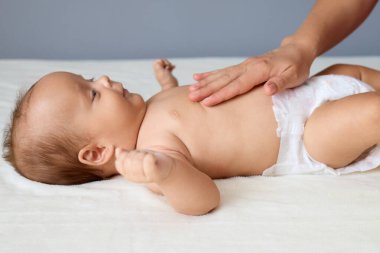 Meçhul kadın, yeni doğmuş sağlıklı beyaz bebeğine nazikçe masaj yapıyor. Beyaz çarşafın üzerinde bebek bezi takıyor. Anne, karın ağrısını azaltmak için karnını rahatlatıyor.
