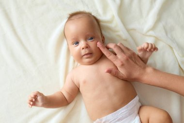 Sevimli, mutlu, yumuşak bebek bezi takan, beyaz çarşafta yatan, tanınmayan bir anne. Sevecen bir şekilde küçük yanağına krem sürerek çocuğun rahat etmesini ve rahatlamasını sağlıyor.