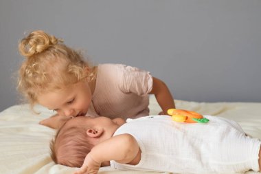 Kıvırcık saçlı, günlük kıyafetler giyen küçük bir kız. Ağlayan erkek kardeşini nazikçe öperek rahatlatıyor. Ailesinin samimi yatak odasında, ablasının evde bebekle tanışması için yatakta oturuyor.