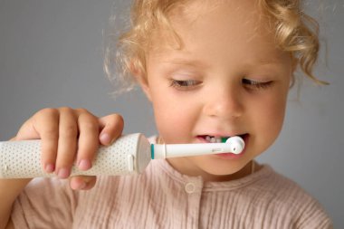 Tatlı komik çocuk dişlerini diş fırçası ve diş macunuyla temizliyor günlük diş bakımının keyfini çıkarıyor ve gri arka planda izole edilmiş bir şekilde diş bakımına odaklanıyor.