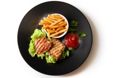 Kızarmış pirzola ya da ızgara et yeşil spatula yaprakları, patates kızartması ve sosu beyaz arka planda izole edilmiş siyah tabakta bulunur.