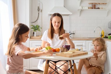 Küçük kızlarıyla birlikte masada taze yemek yiyen mutlu bir kadın evde yemek yerken ve açık mutfakta iletişim kurarken eğlenceli ve sıcak ebeveynlik ve aşk anları yaşıyor.