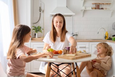 Neşeli bir beyaz anne ve sevimli çocukları mutfakta sağlıklı bir yemek hazırlıyorlar evde birlikte yemek pişirirken mutlu ve sevgi dolu bir ortam yaratıyorlar.