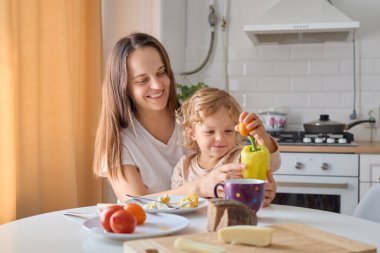 Gülümseyen bir anne, çocuğunu yemek masasında mutlu aile yemeği sırasında sağlıklı sebzelerle besliyor. Küçük kız, sevgi dolu annesiyle lezzetli bir kahvaltı yapıyor.