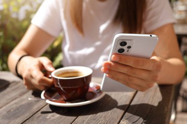Tanımlanamayan bir kadın elinde cep telefonu ve kahve fincanı tutarken internet sayfasında geziniyor güneşli bir yaz gününde kafede kahve molası veriyor ve mesajlaşıyor.