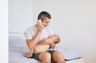 Yeni doğmuş oğlu olan baba, bebeğini kucağında tutuyor ve sohbet ediyor. Akıllı telefonu cep telefonuyla cep telefonuyla açık yatak odasında konuşuyor..