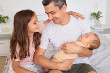 Yeni doğmuş bebeğini sıcak yatak odasında tutarken gülümseyen bir baba taşıyarak aile yaşamlarının neşeli atmosferinin tadını çıkararak şefkat ve mutluluğu ifade ediyor.