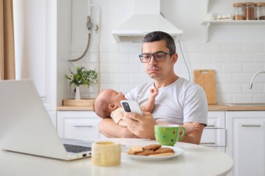 Üzgün, çekici genç bir adam, dizüstü bilgisayarda çalışan genç bir baba, mutfakta bebeğe bakarken akıllı telefondan iş e-postalarını karıştırıyor, uzaktan işleri ve ebeveynliği dengeliyor.