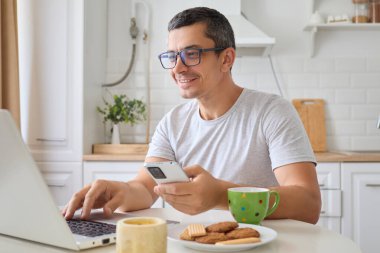 Evden uzakta cep telefonu ve dizüstü bilgisayarla internet ve web kaynaklarına bağlı olarak iş ve teknoloji projeleriyle uzaktan yakından ilgilenen neşeli, serbest giyinen bir erkek.