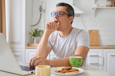 Evde dizüstü bilgisayar kullanarak çalışan yorgun bir adam kahve ve kurabiyeyle masada otururken kablosuz internet bağlantısı kullanıyor. Uzaktan çalışma sırasında yorgun ve esniyor.