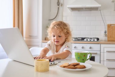 Küçücük dalgalı saçlı küçük kız mutfak masasında dizüstü bilgisayar oyunu oynuyor. Teknolojiyi kullanıyor. Bu arada anne de internetteki karikatürleri açmaya çalışıyor.