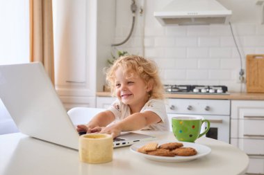Kıvırcık saçlı kız çocuk, çizgi film seyrediyor. Gülümsüyor. Mutfaktaki masada oturuyor.