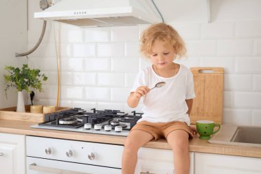 Sevimli çocuk mutfak tezgahının tepesinde oturuyor ve kaşık küçük çocuğa bakıyor kıvırcık sarı saçlı ve beyaz tişörtlü kahvaltısını bekliyor.